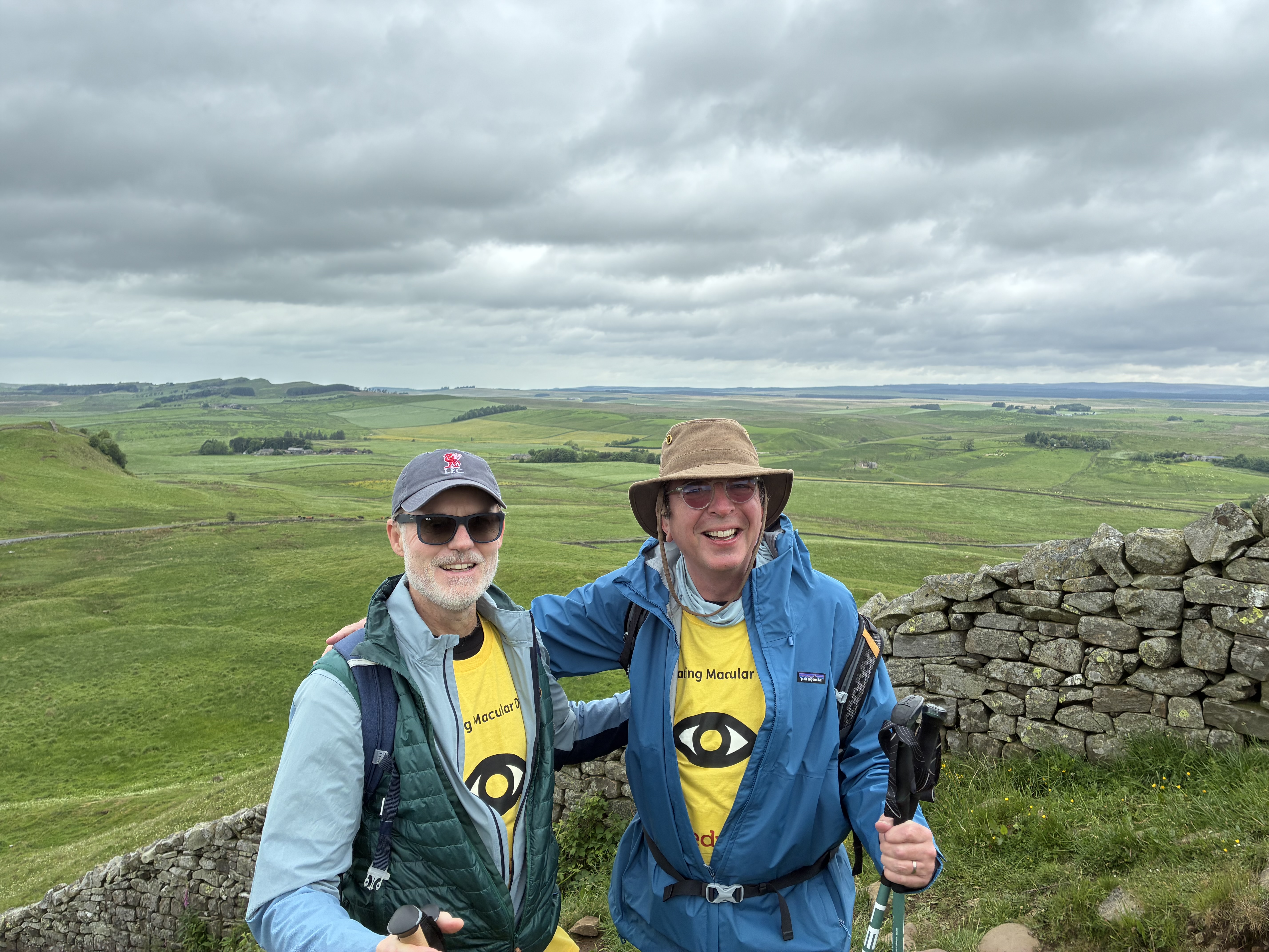 David and Tord in macular tshirts and wearing walking gear, smiling at the camera with a view of a large field and rolling hills in the background 