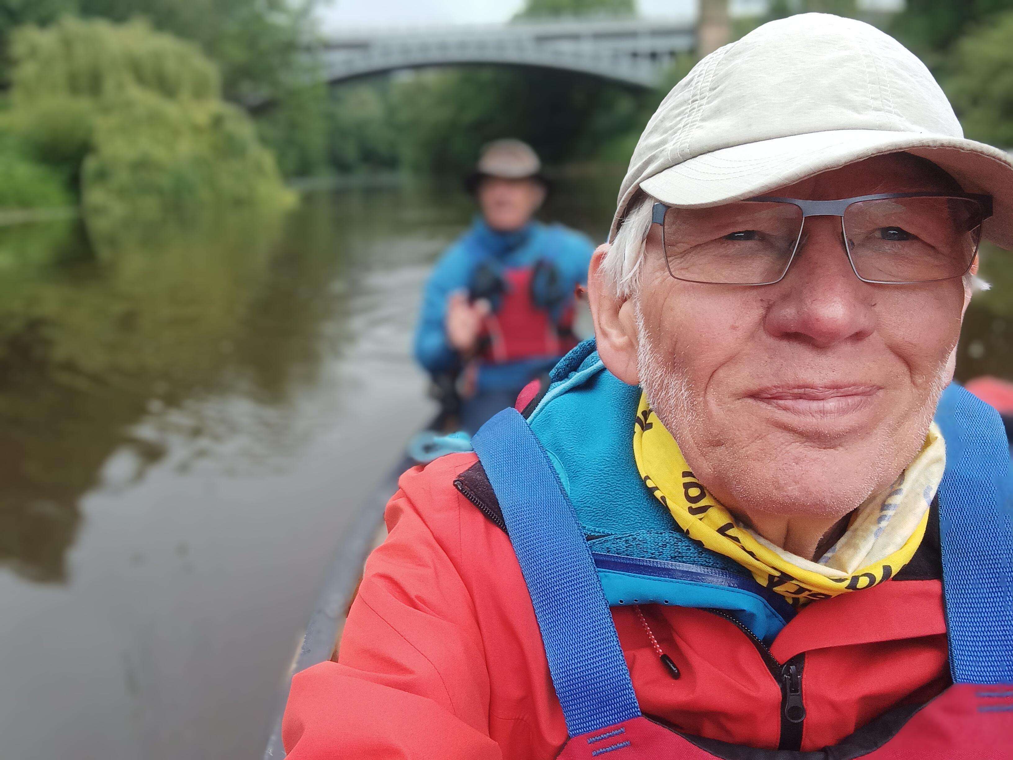 David in the canoe wearing hat and glasses and smiling to camera, with blurred outline of Norman in the background