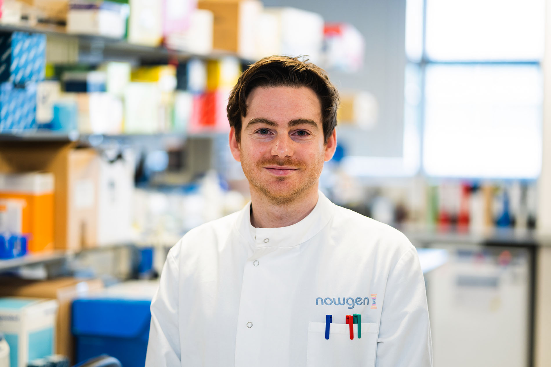 Jamie Ellingford, a white male researcher with brown hair posing in a lab coat in the lab, smiling to camera 