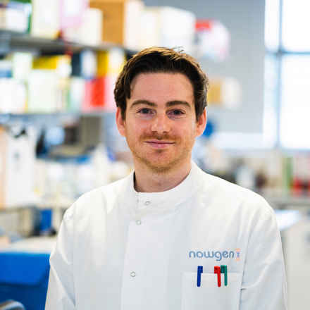 Jamie Ellingford, a white male researcher with brown hair posing in a lab coat in the lab, smiling to camera 