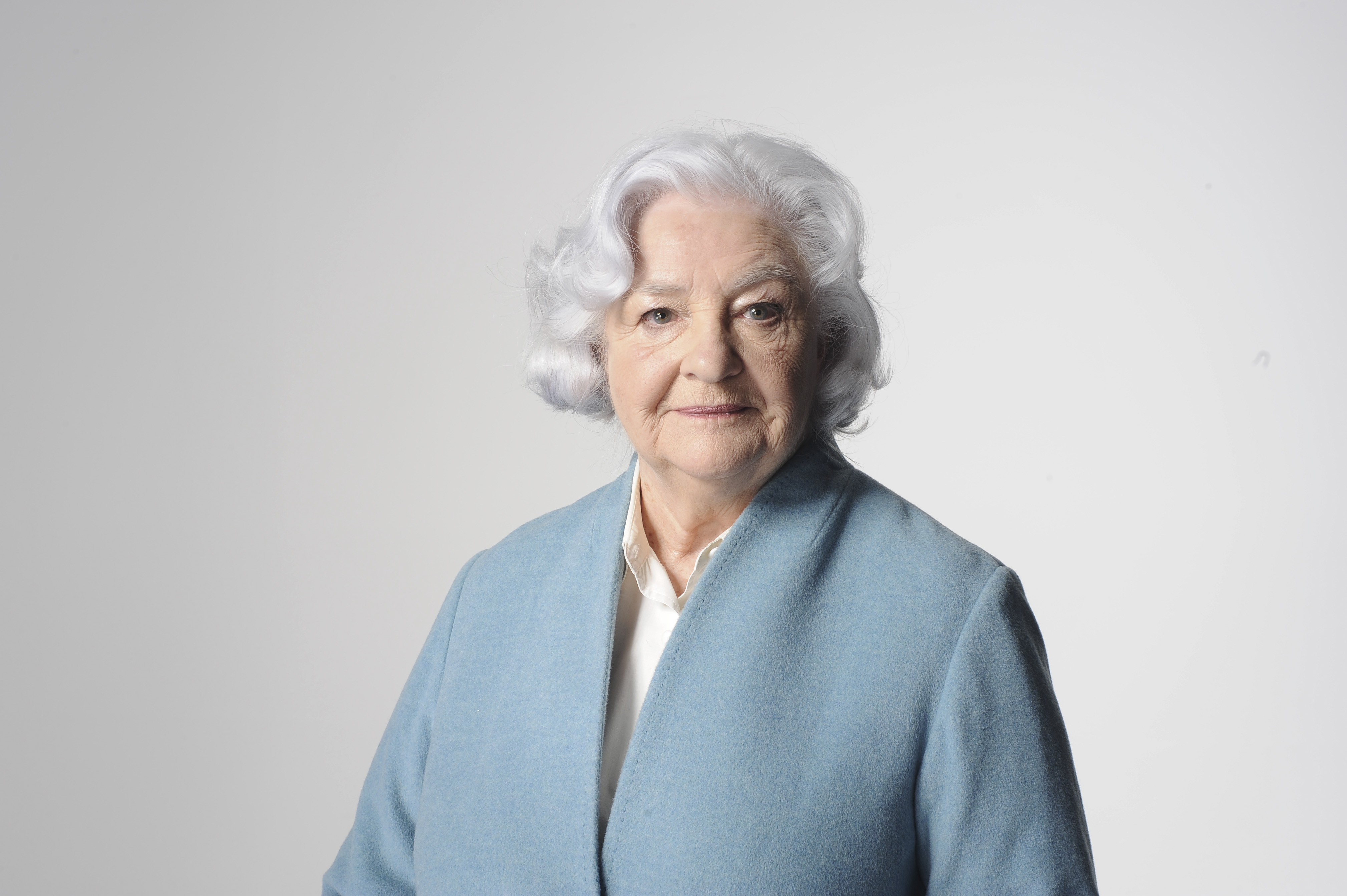 Patricia Greene, a woman in her 80s, with short grey hair, looking towards camera in light blue jacket with plain background