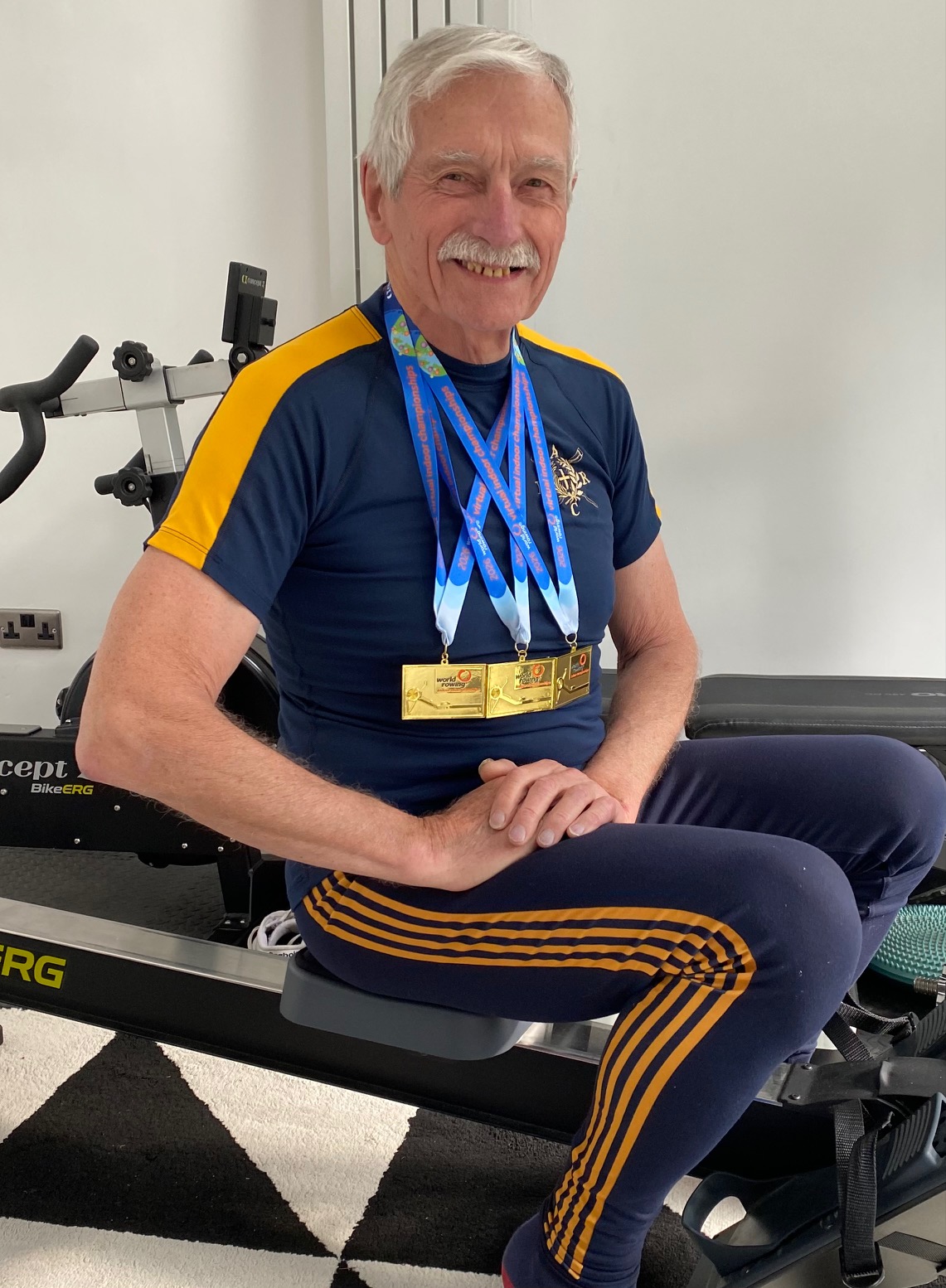 Roger, 81-year old white man in gym kit, sat on an indoor rowing machine and smiling to camera. He is wearing his medals. 