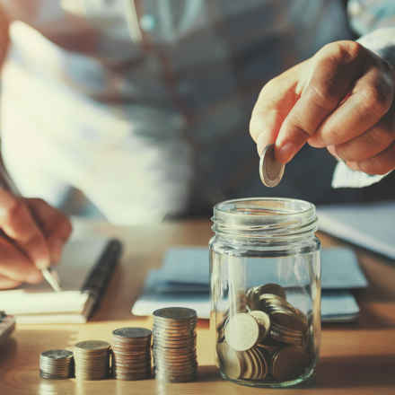 A man counting his money, while writing down calculations. There are four piles of coins, each growing in size, next to a jar of coins.