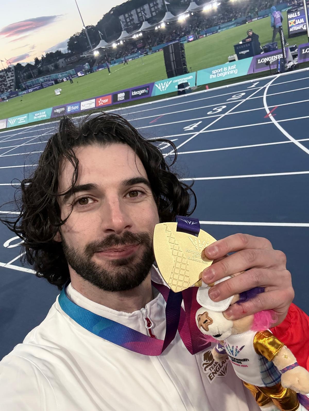 Zac, a white man in his 30s with dark brown hair and a beard, smiling and holding his gold medal to camera 
